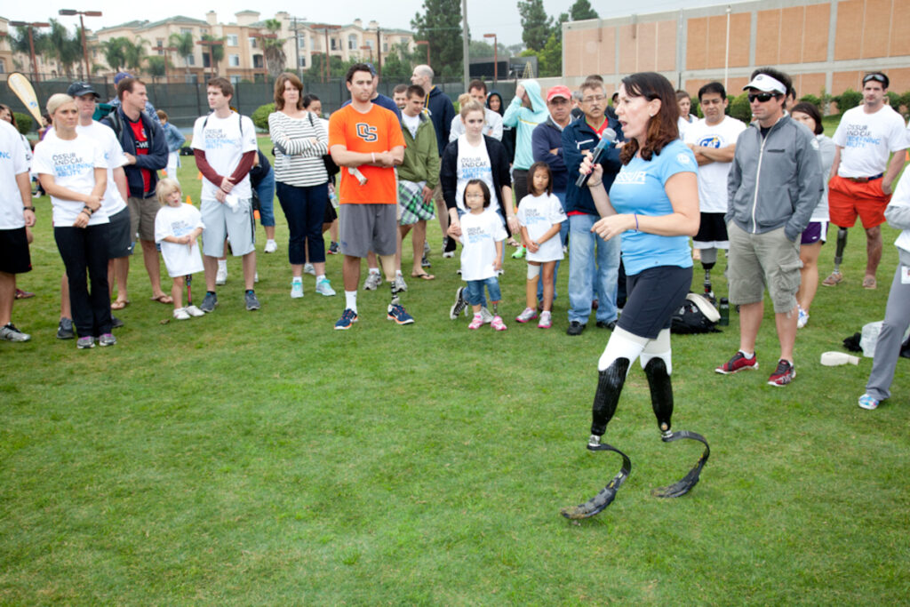 A person with prosthetic legs speaks to an attentive group outdoors on a lawn.
