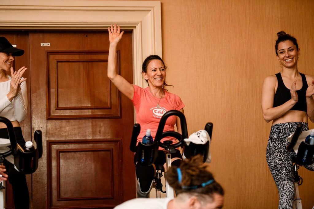 A person in a pink shirt raises her hand while sitting on a stationary bike, with others clapping nearby.