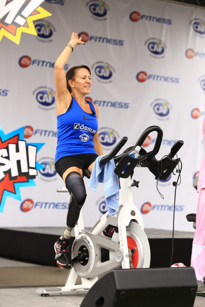 A person with a prosthetic leg exercises on a stationary bike, raising one arm under a banner with fitness logos.