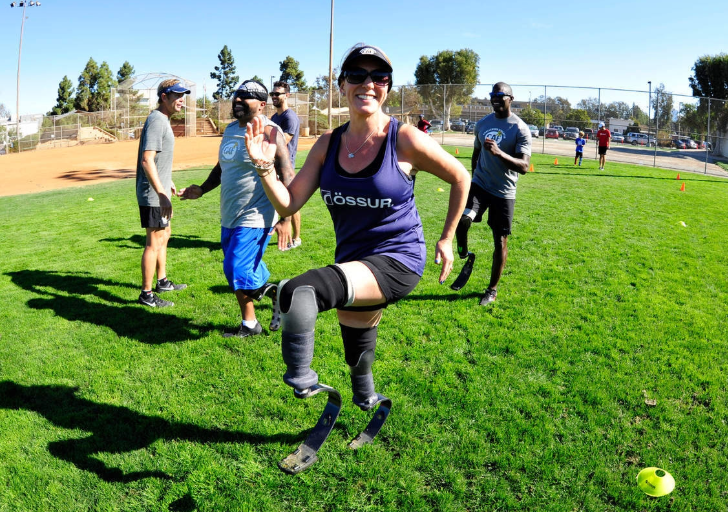 A woman with prosthetic running blades running on a grassy field, with a group of people watching.
