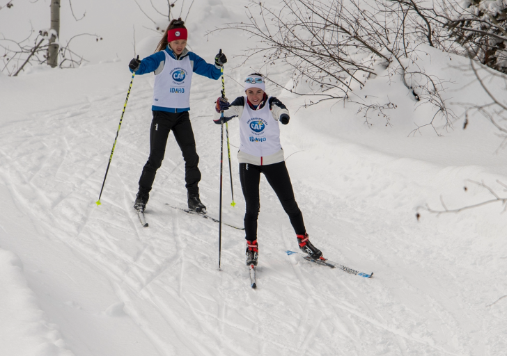 Two cross-country skiers on a snowy trail wearing "CAF IDAHO" vests.
