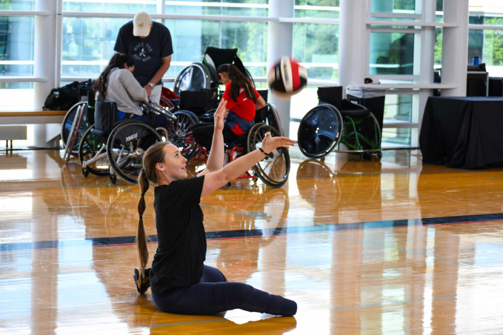 A person with a lower limb difference sitting on a gym floor, reaching to hit a volleyball, with people in wheelchairs in the background.