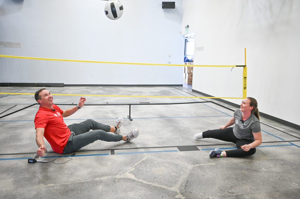 Two people sitting on a floor playing with a ball over a low net indoors.