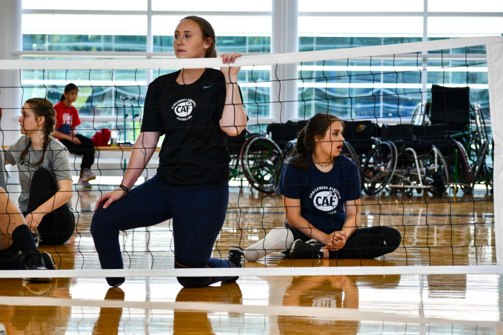 Participants in a seated volleyball game wearing "CAF Challenged Athletes Foundation" t-shirts in a sports facility.