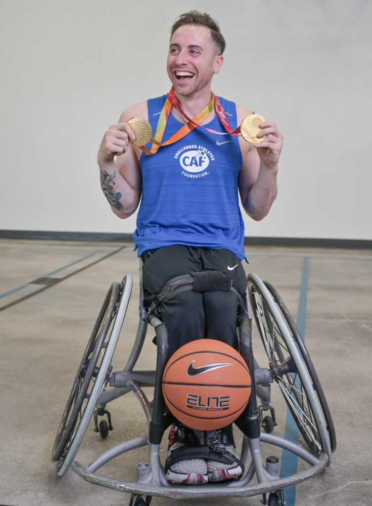 A smiling person in a sports wheelchair wearing a blue athletic shirt with two gold medals and a basketball in front.
