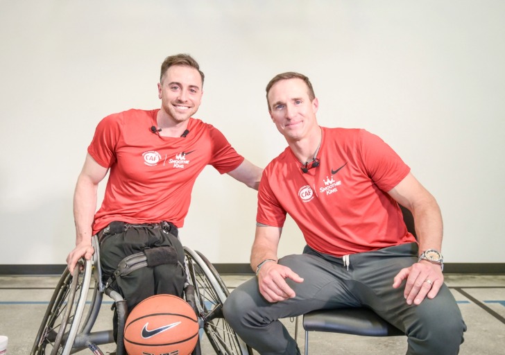 Two people in red shirts, one in a wheelchair with a basketball, smiling at the camera.