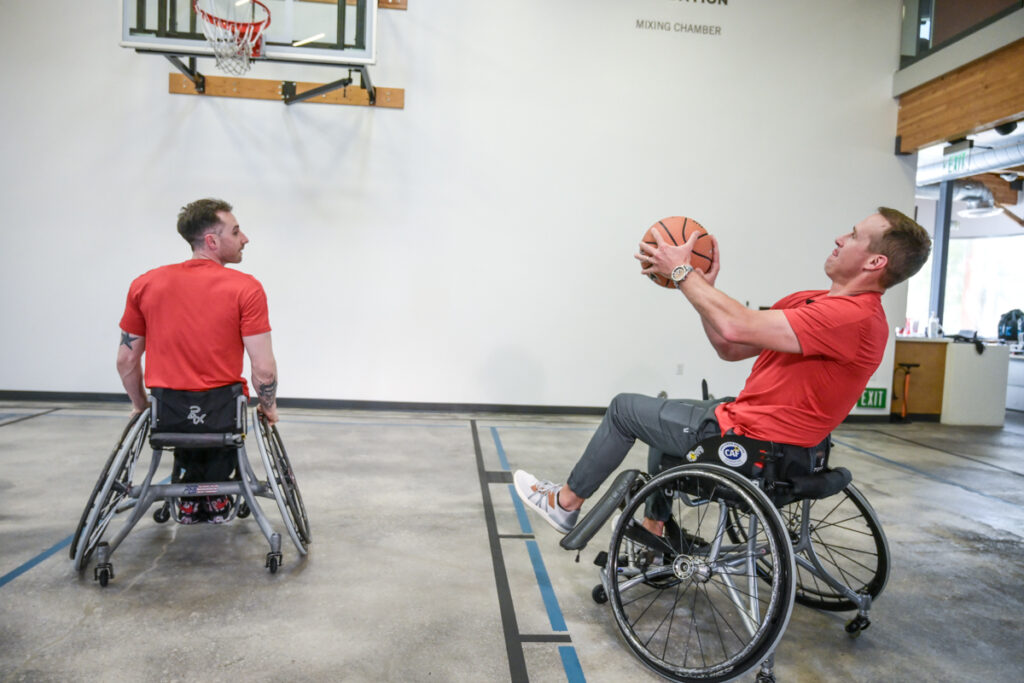 Two men in wheelchairs playing basketball indoors, one preparing to shoot.