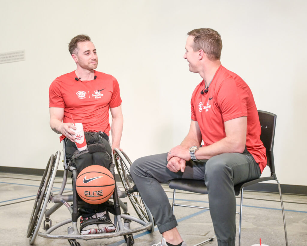 Two men, one in a wheelchair with a basketball, talking indoors.