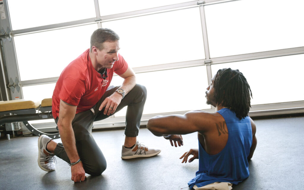 A person in a red shirt kneeling and talking to another person on the gym floor.