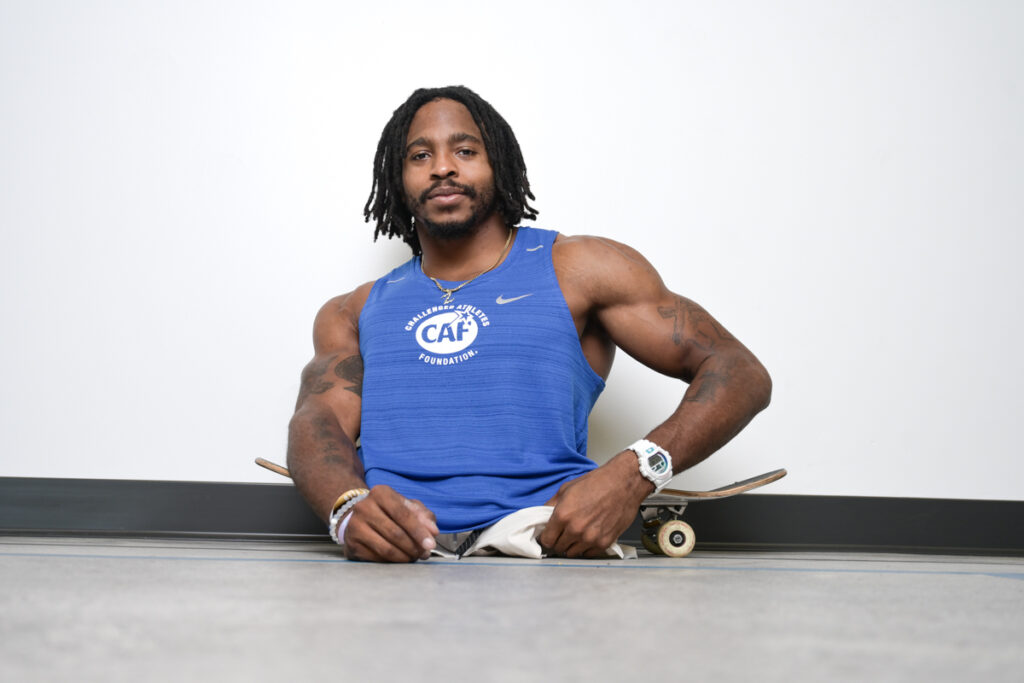 A muscular person without legs sits near a skateboard, wearing a blue sleeveless shirt with the Challenged Athletes Foundation logo.