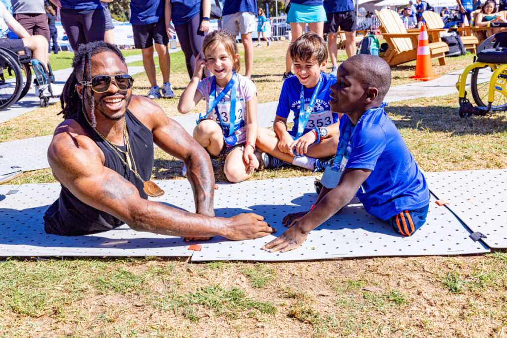 A muscular person in sunglasses interacts with a young child on an exercise mat outdoors, surrounded by other children and event attendees.