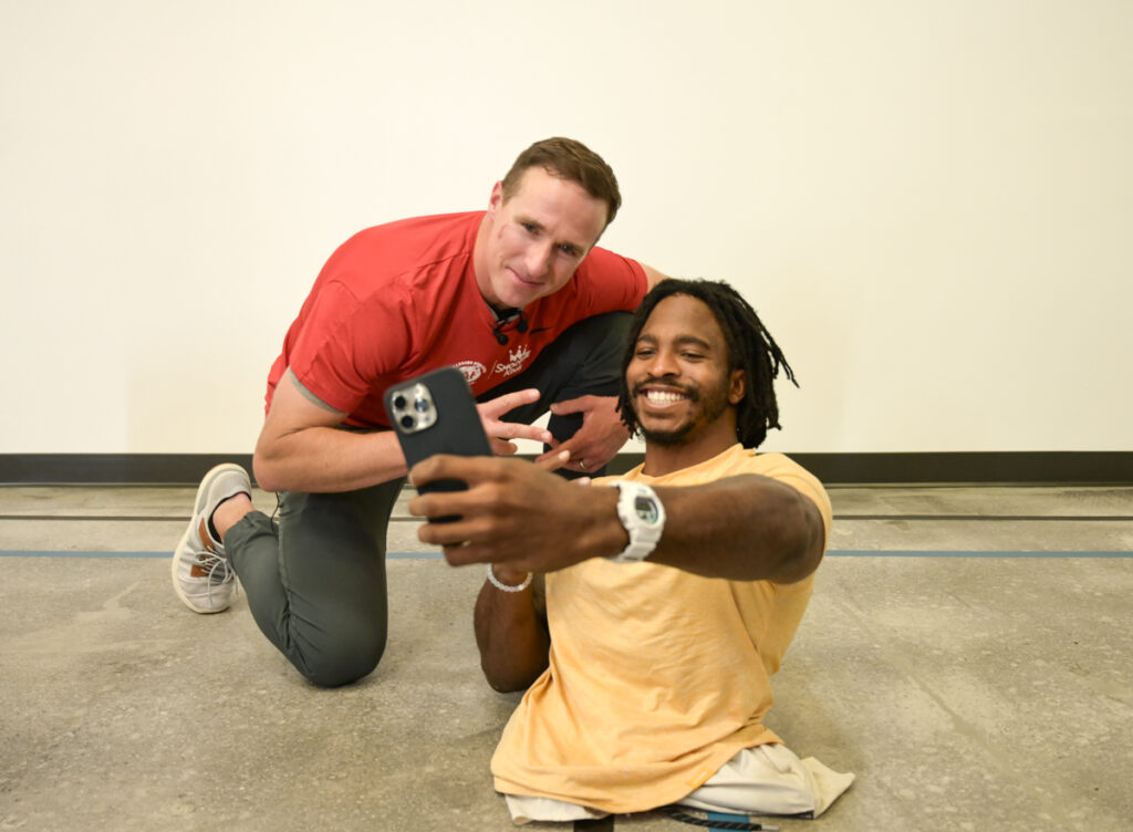 Two people taking a selfie indoors, one of whom has no legs. One is sitting and smiling, the other is giving a peace sign.