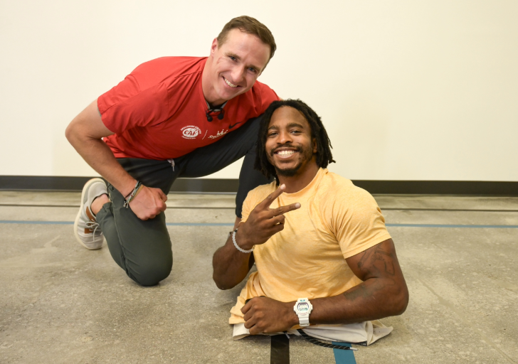 Two people indoors smiling at the camera; one kneeling, the other on the floor.