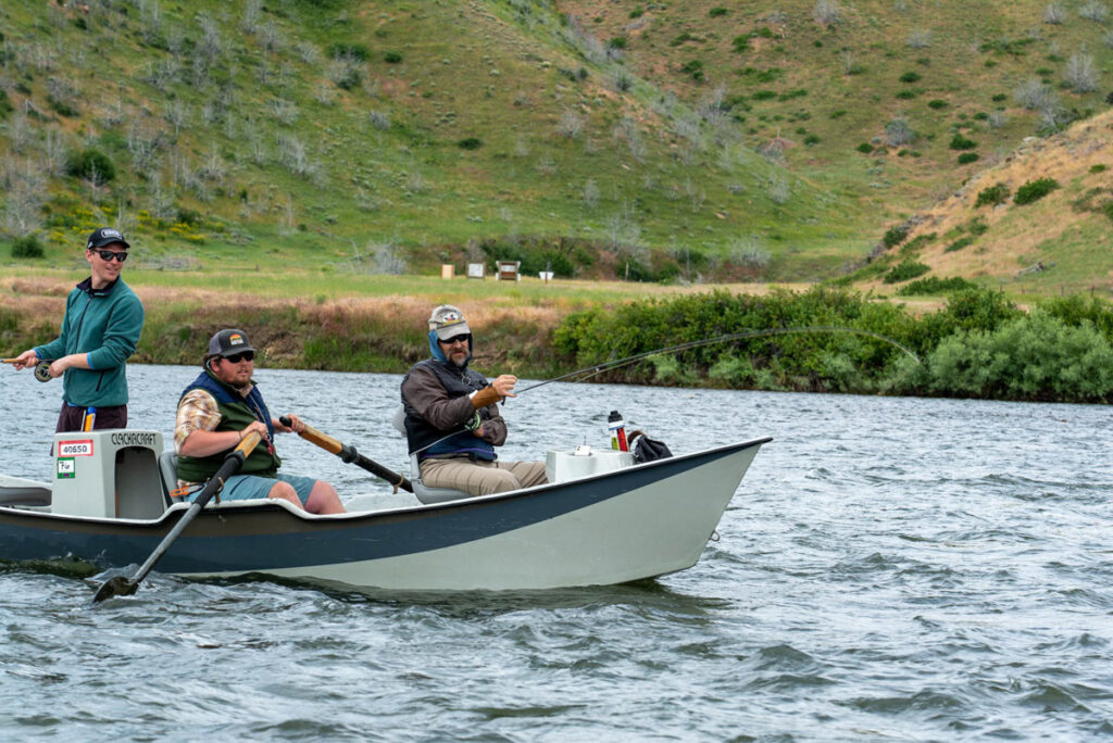 Three men fishing in a boat on a river surrounded by grassy hills.