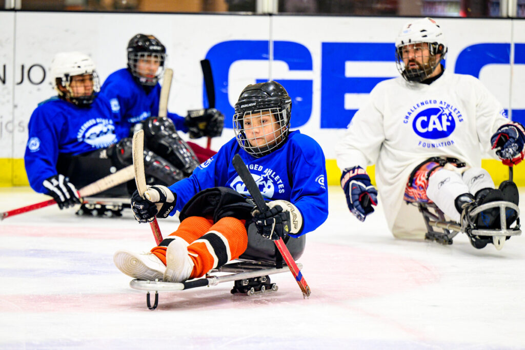 Athletes play sled hockey on an ice rink. A young player in the foreground wearing an orange uniform and blue Challenged Athletes Foundation jersey moves forward using hockey sticks. Other players in blue and white jerseys are visible in the background, also on sleds, wearing helmets and protective gear.
