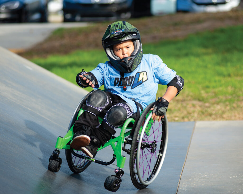 A young athlete in a helmet and protective gear maneuvers a green wheelchair on a concrete ramp. The athlete wears a blue shirt, black pants, and knee pads, focusing intently while leaning into the turn. Grass and parked cars are visible in the background.