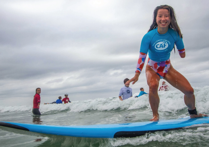 A woman with an upper limb difference in a blue wetsuit surfing on a light blue board, smiling with a group of surfers in the background.