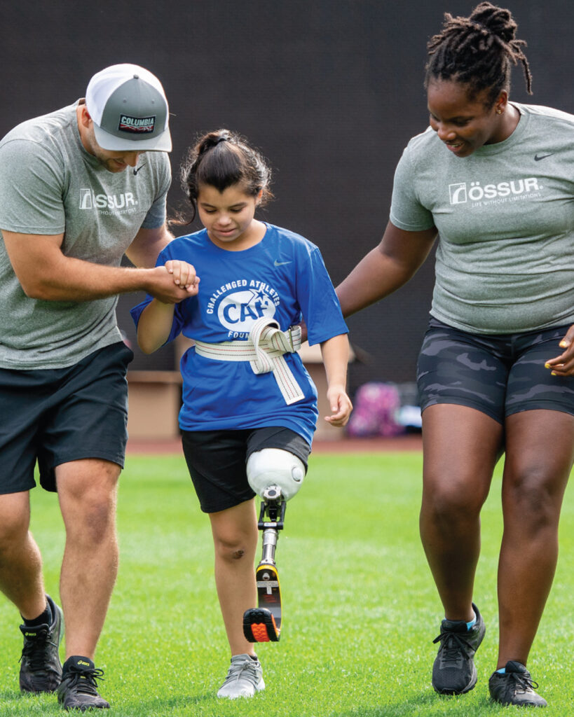 A young girl with a prosthetic leg walks on grass helped by two adults.