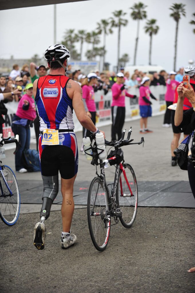 A triathlete with a prosthetic leg holds a bike, surrounded by spectators near palm trees.