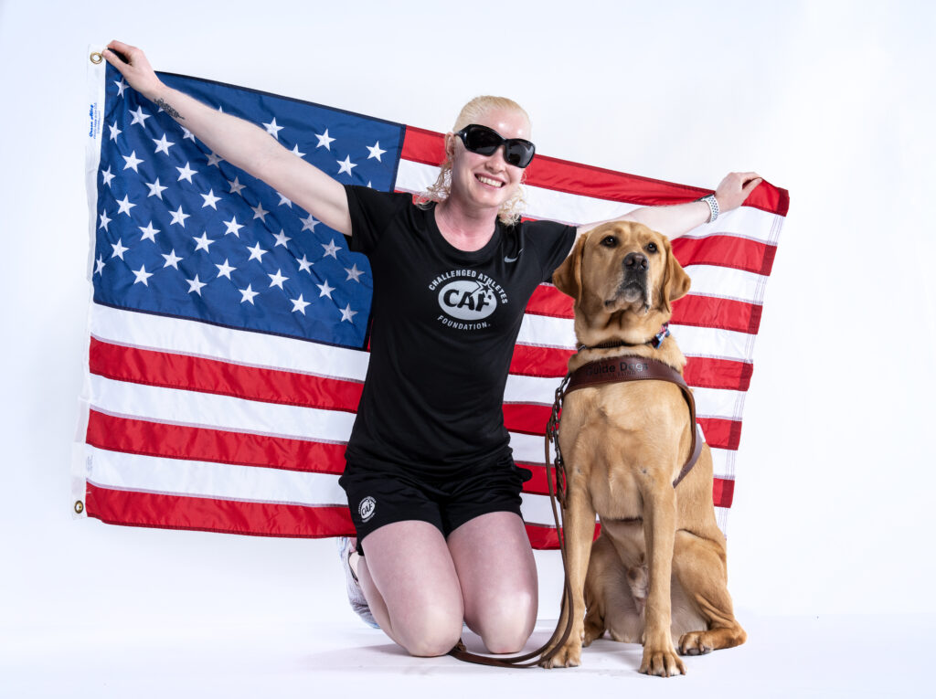 A person wearing sunglasses and a black Challenged Athletes Foundation t-shirt kneels on one knee while holding up an American flag. Beside her sits a guide dog wearing a harness. Both are facing the camera against a plain white background.