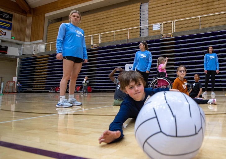 Children playing seated volleyball in a gym with a coach watching.