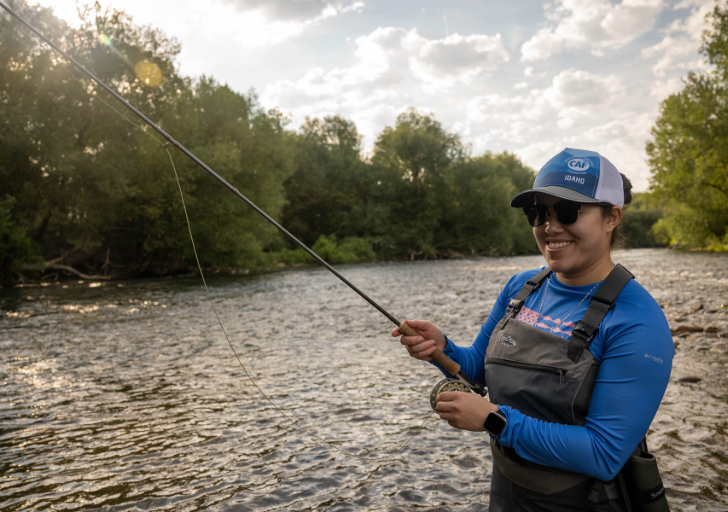 Person fishing in a river with greenery and cloudy sky in the background.