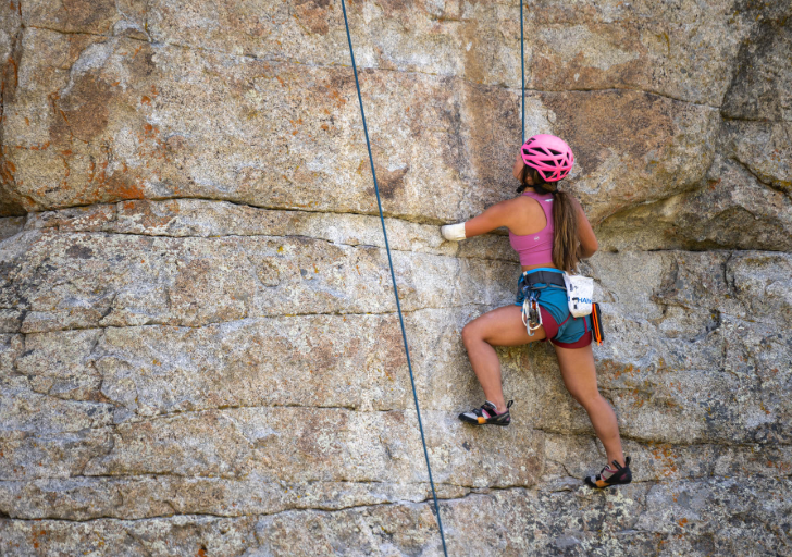 A female rock climber in a pink helmet ascends a rugged rock face.