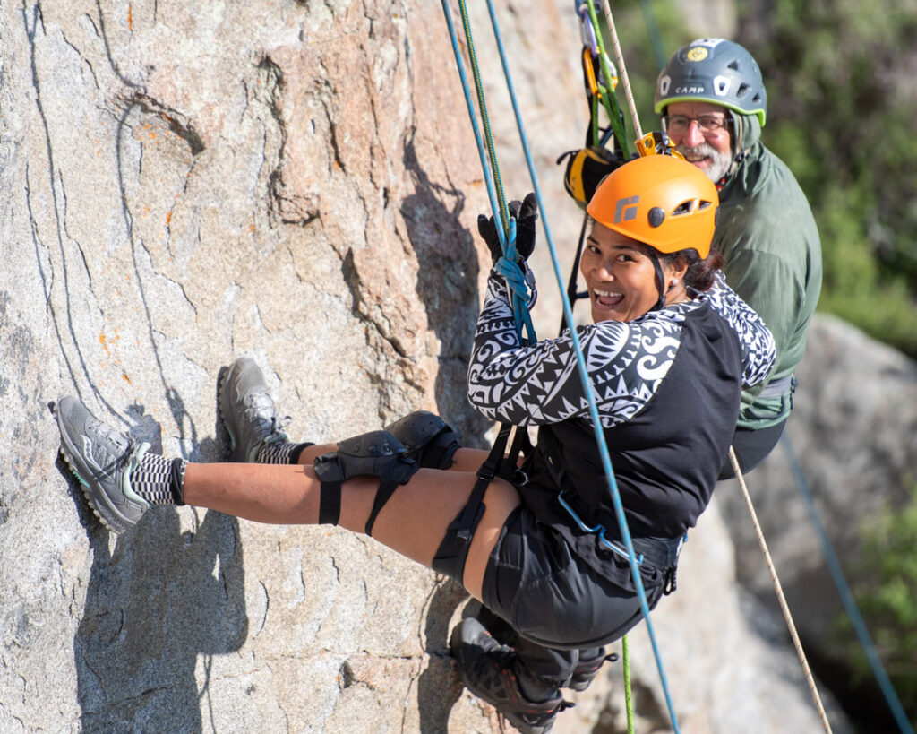 Two climbers, a woman and a man, smiling while ascending a rocky cliff face with ropes and harnesses.