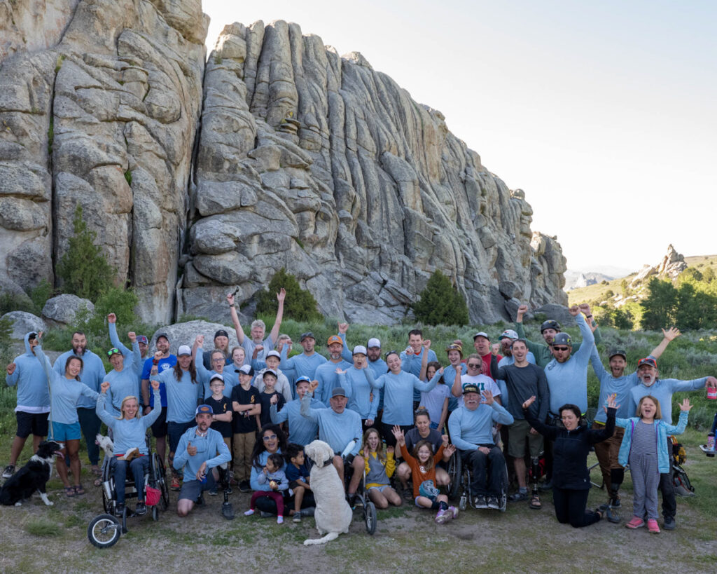 A diverse group of people posed in front of a large, rocky formation, some raising their arms in celebration.