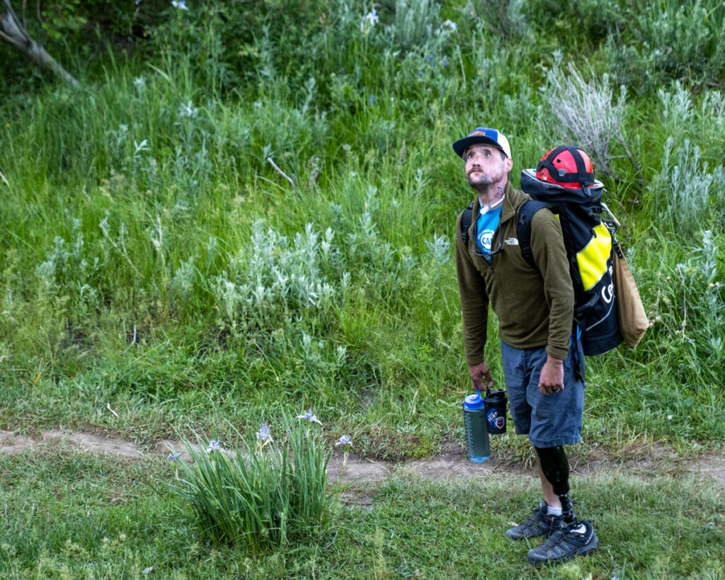 A man with a prosthetic leg is standing on a grassy path with a large backpack, a helmet, and water bottles. Surrounding him are tall grasses and green vegetation.