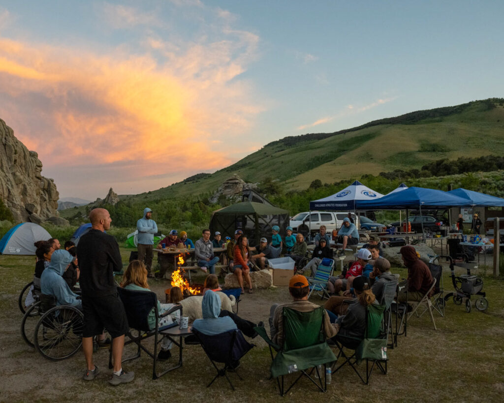 A group of people seated around a campfire at a campsite during sunset with hills in the background.