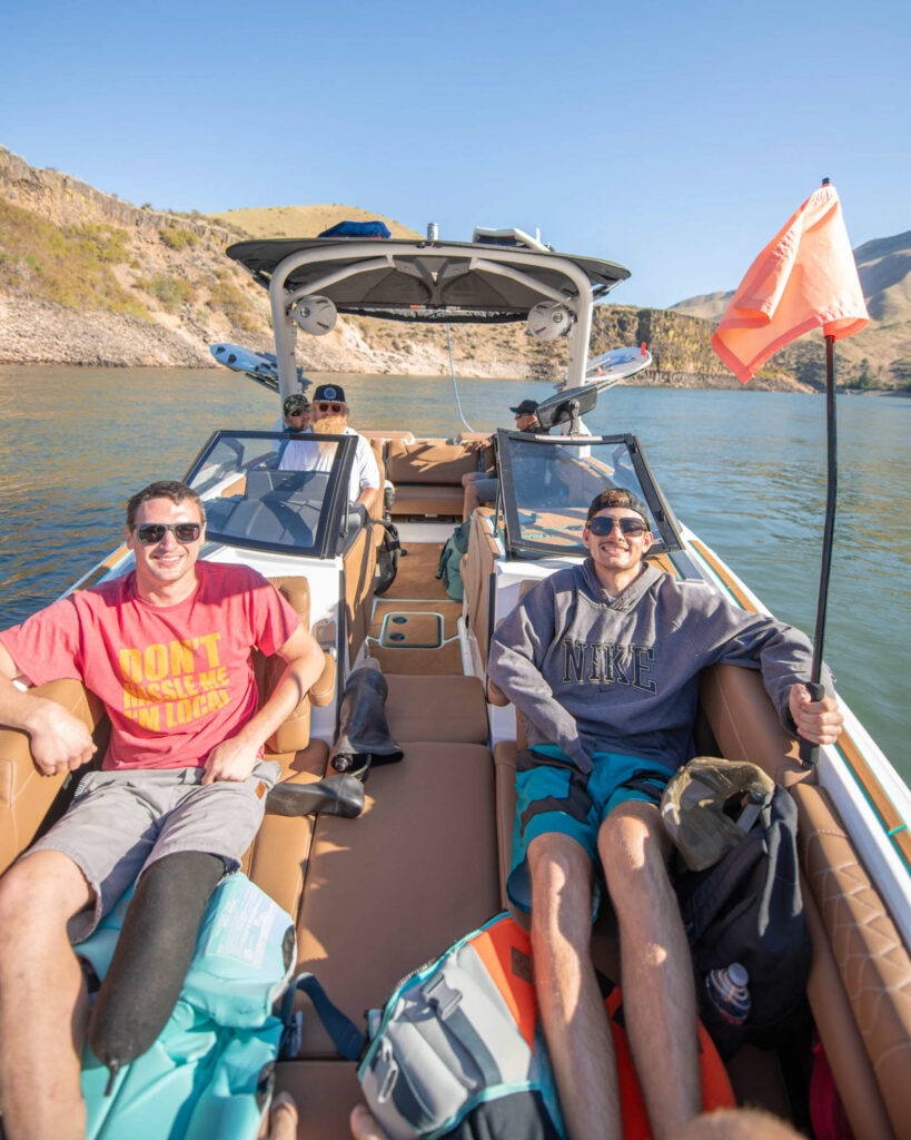 Two men with prosthetic legs seated at the front of a motorboat, smiling on a sunny day.