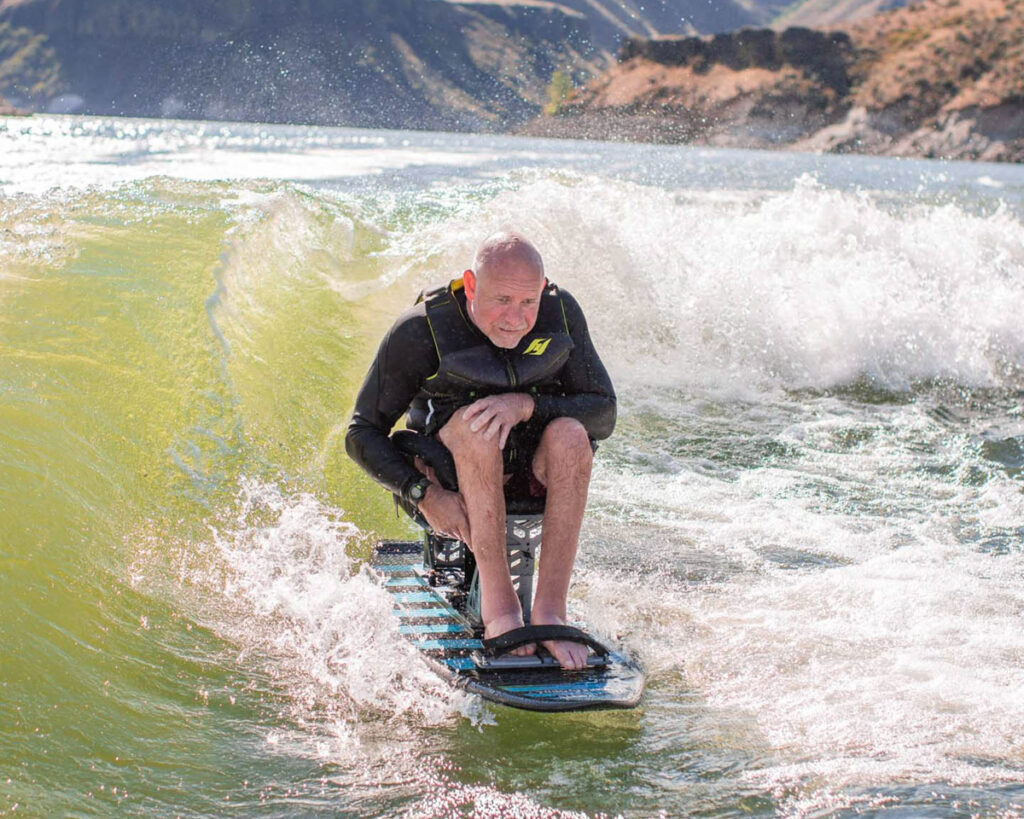 Man in a wetsuit surfing a wave on a striped board with hills in the background.