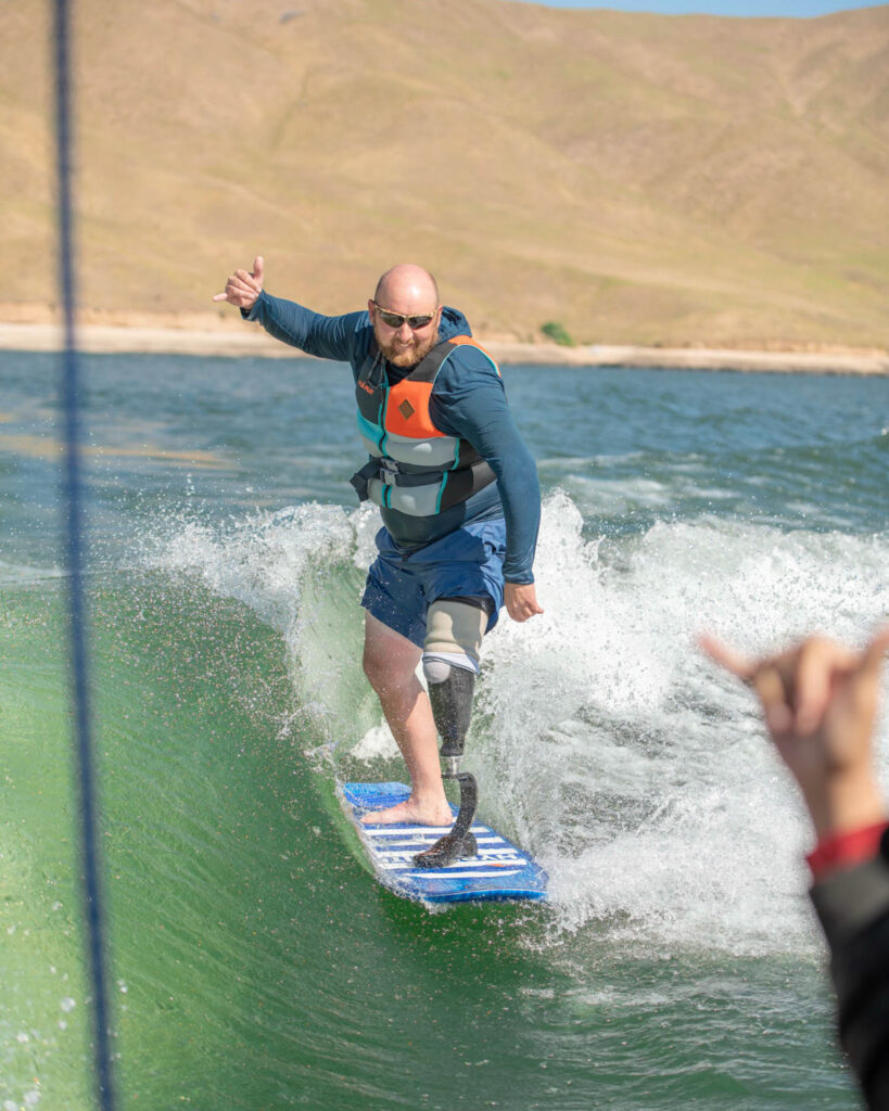 A man with a prosthetic leg surfing on a wave, wearing a blue shirt and life vest, with hills in the background.