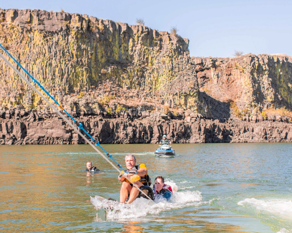 Two people on a water sled being towed near rocky cliffs.