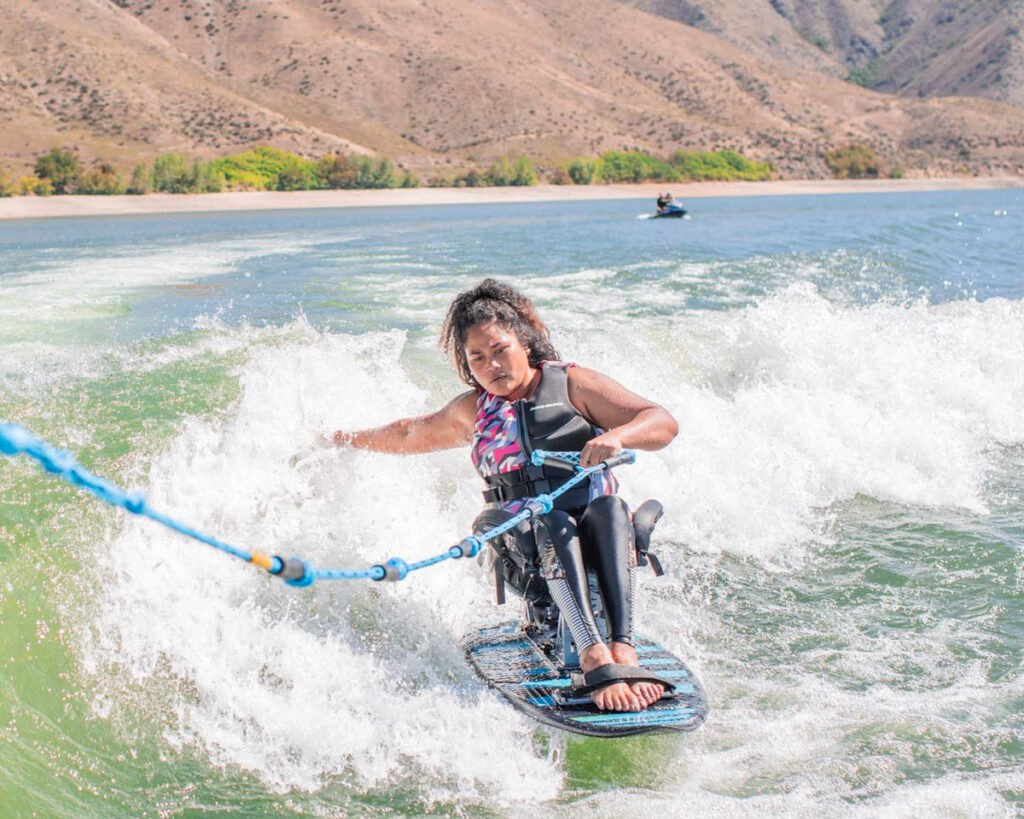 Person wakesurfing with a sit-ski on a lake, holding a blue rope, surrounded by waves.