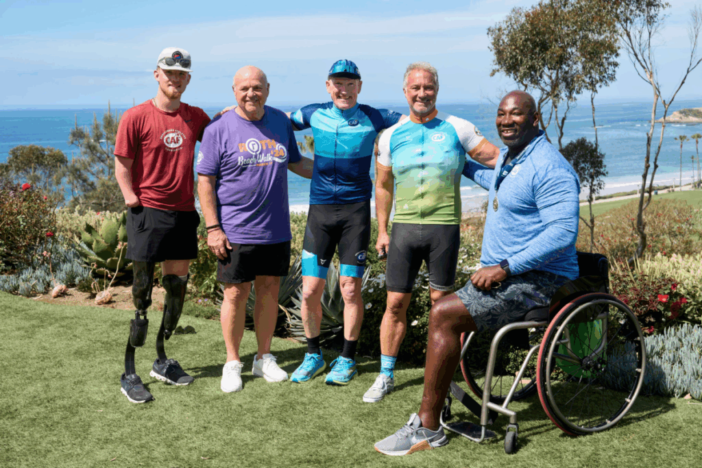 Five men, including athletes with prosthetic legs and a wheelchair user, stand together smiling for a group photo with the ocean in the background.