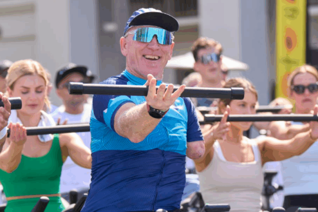 A man with limb difference smiles while leading a cycling session, holding a workout bar, with other riders participating behind him.