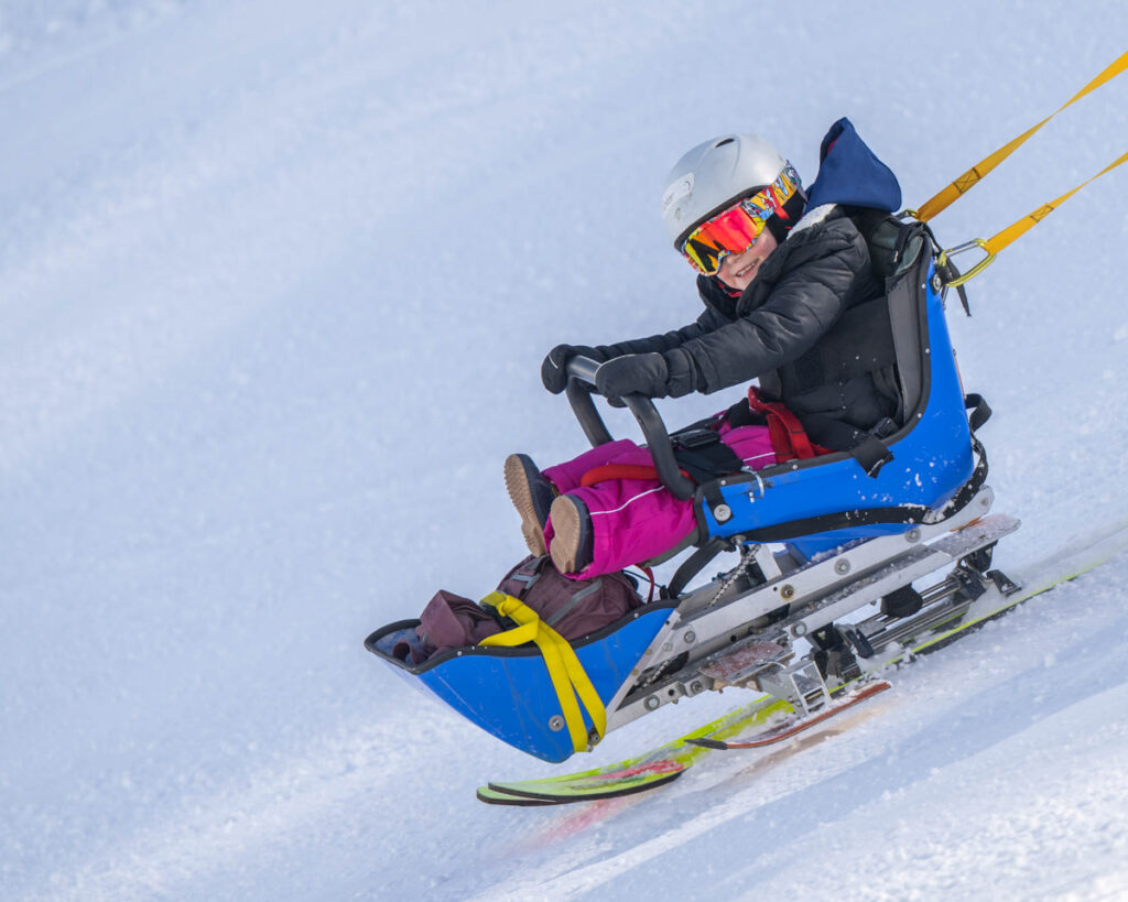 Person using a blue sit-ski on a snowy slope, wearing pink pants and a white helmet.