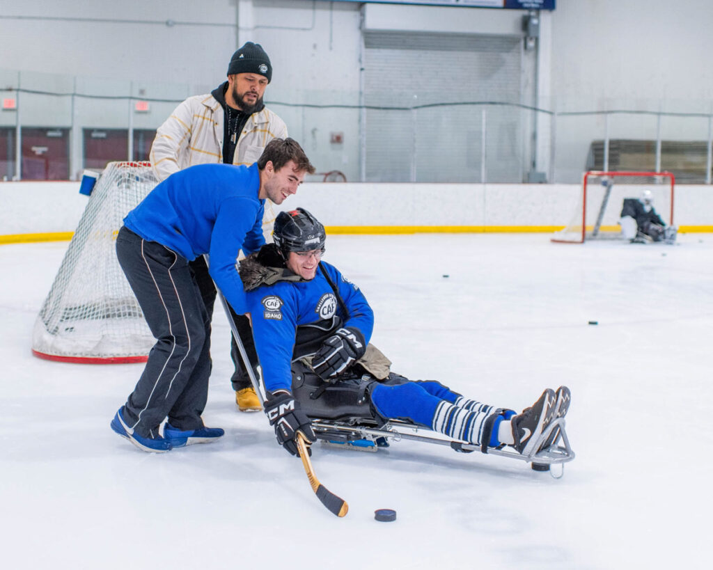Three men on an ice rink, with one in a sled holding a hockey stick while being assisted by the other two.