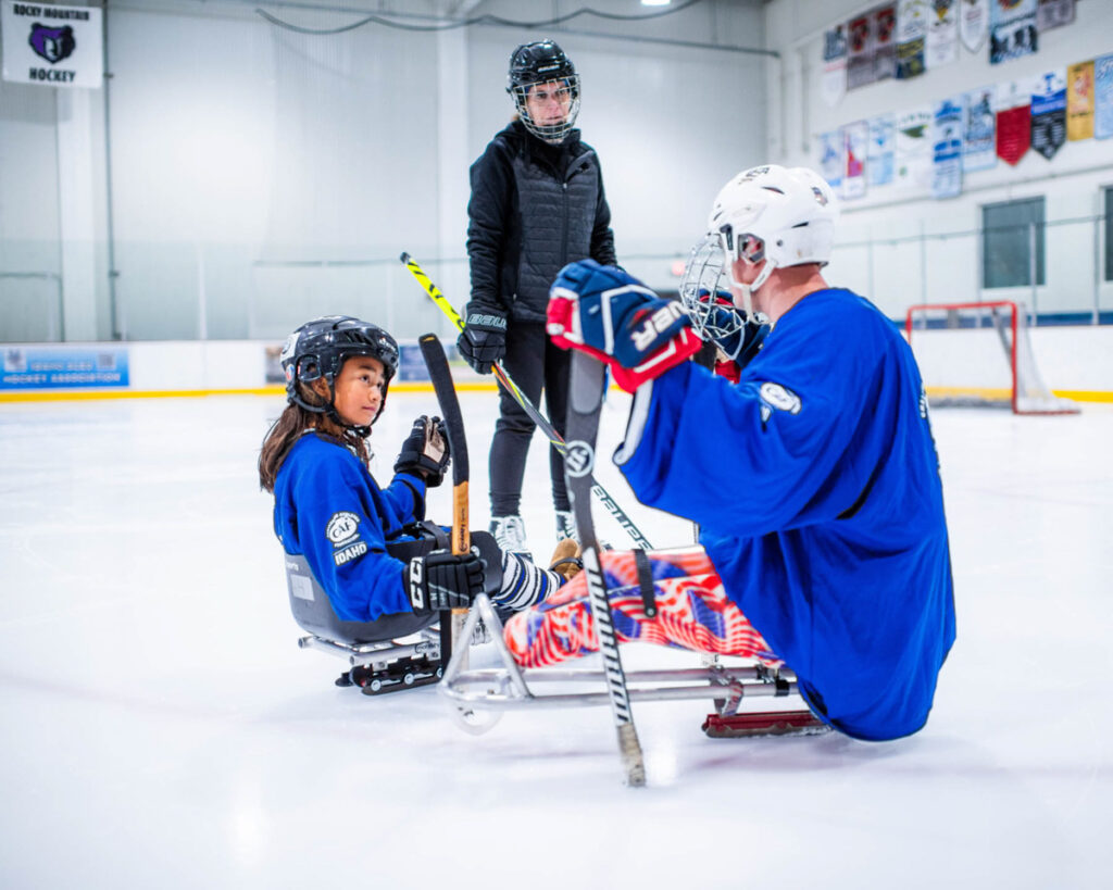 Two people in blue jerseys play sled hockey on an ice rink, observed by a standing person.