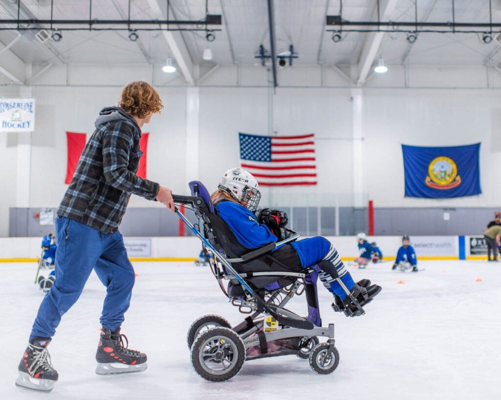 Person in a plaid shirt pushes a wheelchair on an ice rink.