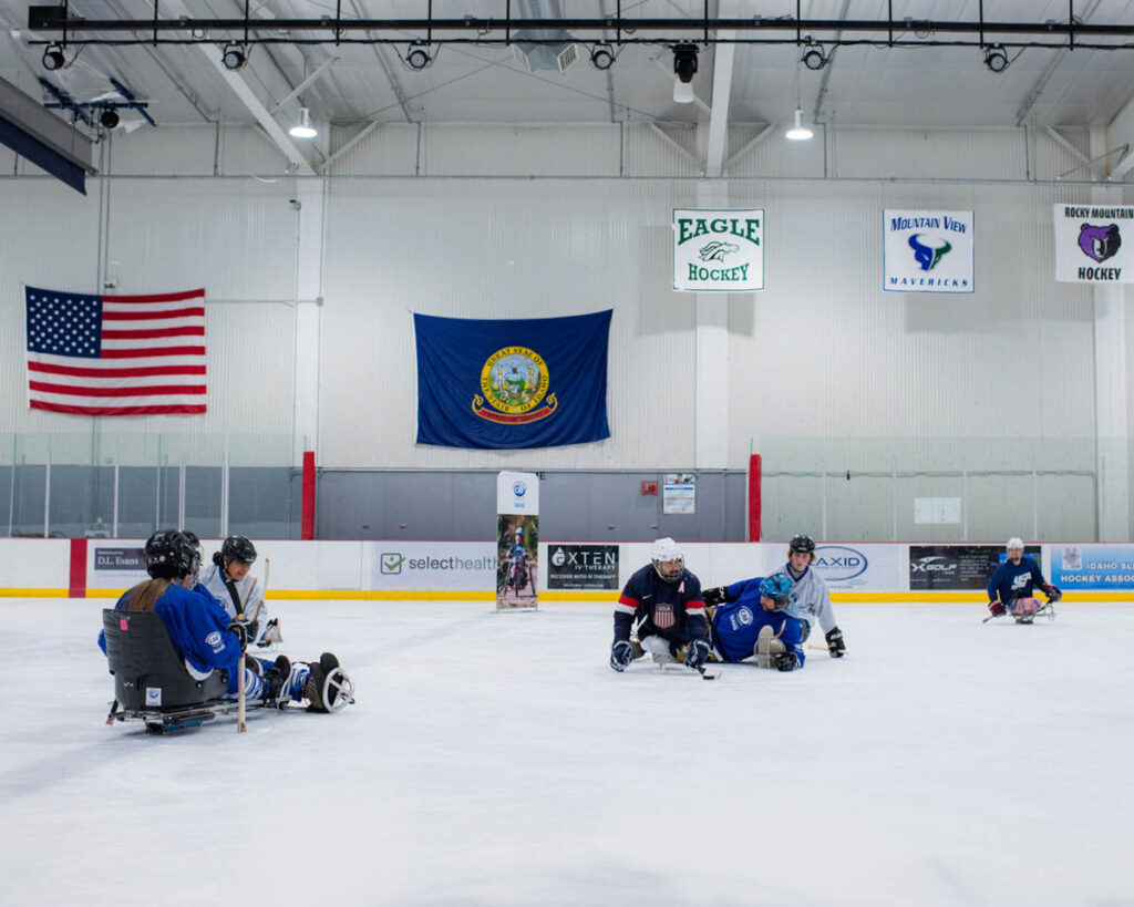 Players engage in a sled hockey game on an ice rink with flags and banners in the background.