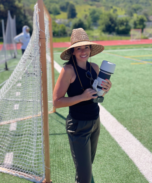 Photographer wearing a sunhat stands on a sports field holding a large camera lens, smiling while standing next to a soccer goalpost