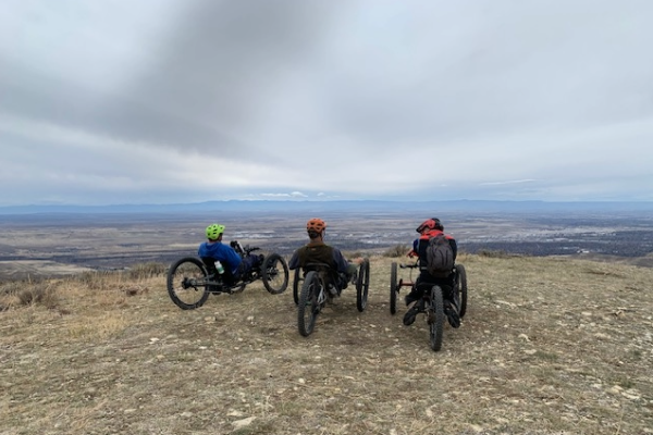 Three adaptive mountain bikers with helmets and backpacks sit on rugged off-road handcycles overlooking a wide valley and distant mountains beneath a cloudy sky.