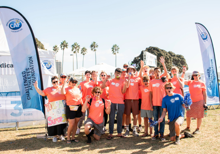 Group of volunteers wearing bright orange shirts posing together with CAF flags at an outdoor event. They smile, cheer, and hold supportive signs with palm trees in the background