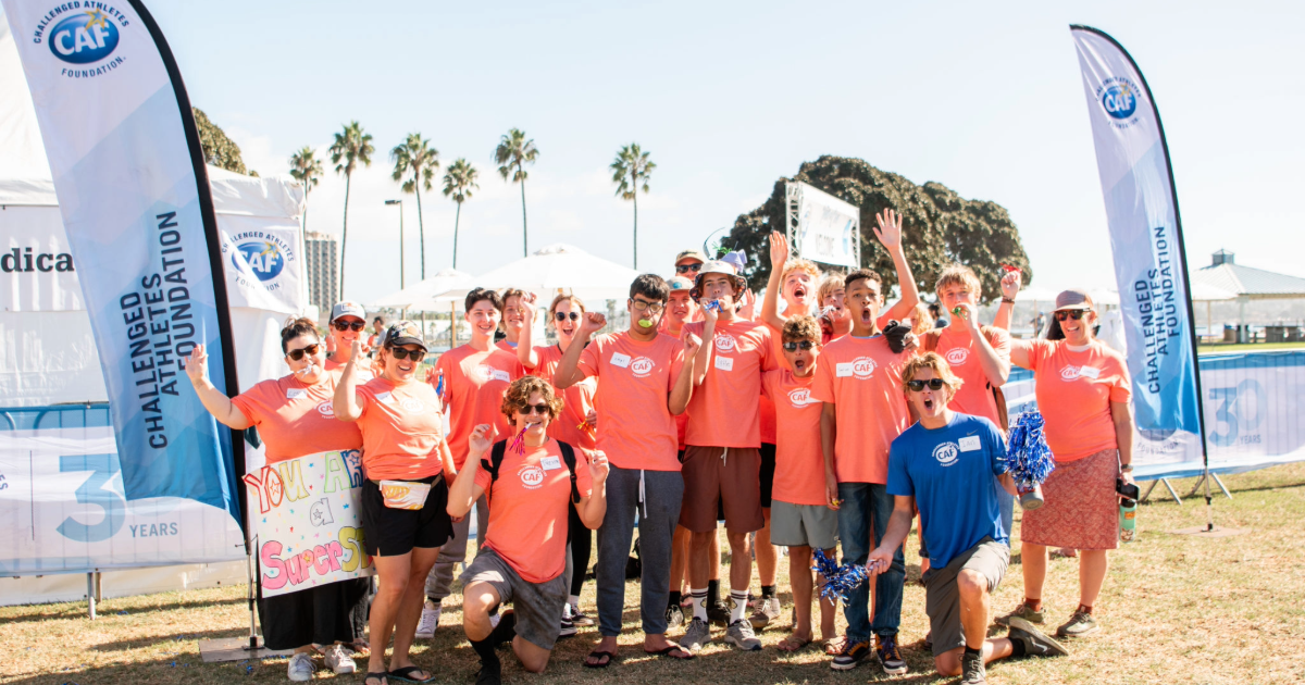 Group of volunteers wearing bright orange shirts posing together with CAF flags at an outdoor event. They smile, cheer, and hold supportive signs with palm trees in the background
