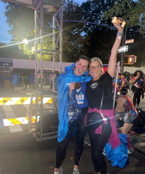 Two women celebrate at the finish line of a race. One wears a medal and a blue finisher’s poncho, while the other raises an arm triumphantly with a big smile.