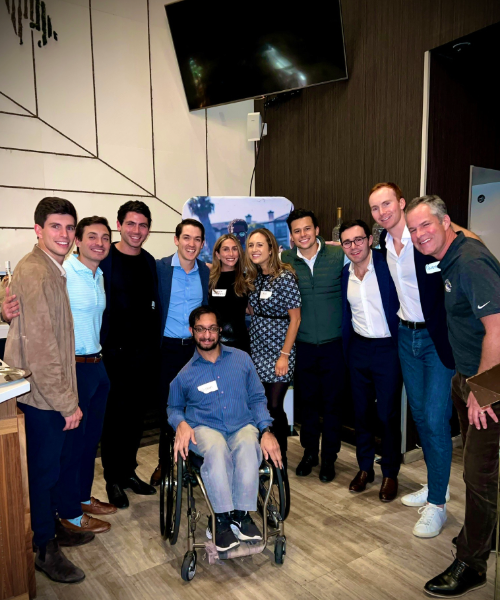 Group of people gathered indoors, smiling together for a photo. One man at the front uses a wheelchair. Everyone appears relaxed and happy, dressed in casual and business attire