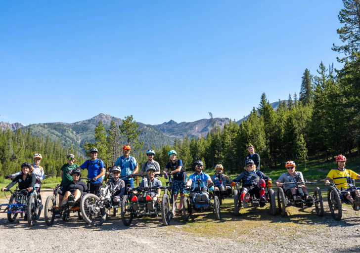 Group of sixteen people posing for the camera while wearing biking gear in the mountains; most of them are on adaptive mountain bikes.